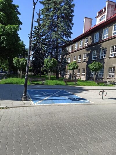 Niebieskie miejsce parkingowe, po lewej latarnia uliczna droga i chodnik z kostki brukowej. Na wprost ławki i skwer z drzewami i trawnikiem. Po prawej szary budynek edukacyjny.