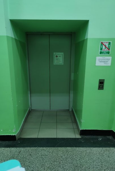 Drzwi zewnętrzne do windy umieszczone we wnęce. Panel przywoławczy windę umieszczony przed wnęką. Nad panelem są dwie tabliczki. Ściana na dole ma kolor ciemno zielony, wyżej jasnozielony.
