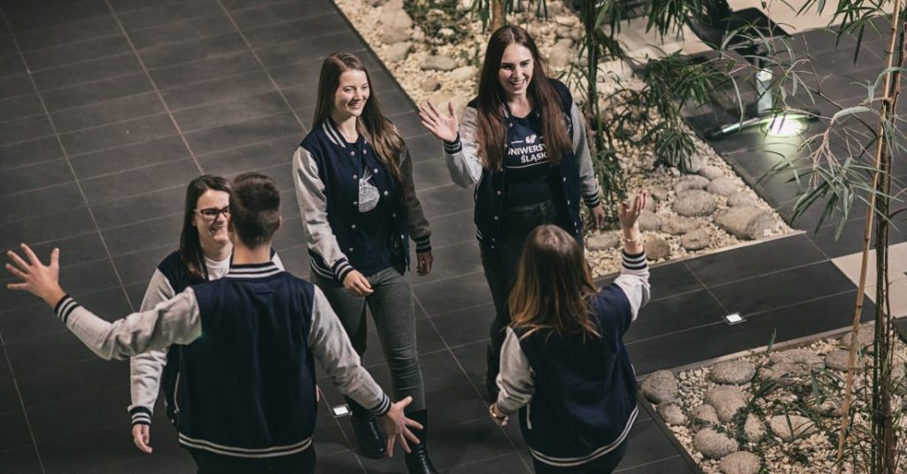 Grupa młodych osób ubranych w uniwersyteckie bluzy wita się radośnie