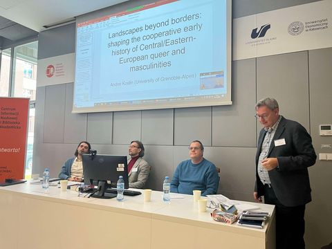 panel dyskusyjny, trzy osoby siedzą przy stole konferencyjnym, czwarta stoi, za nimi ekran rzutnika z wyświetlonym slajdem