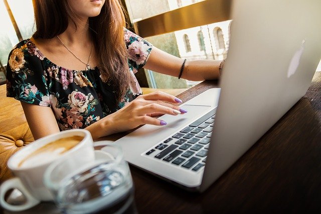 kolorowe zdjęcie: dziewczyna, której nie iwdać twarzy, siedzi przed laptopem. Obok stoi filiżanka z kawą
