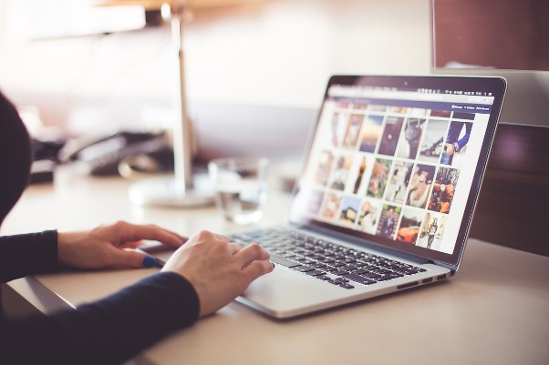 otwarty laptop, na którego monitorze wyświetlają się różne kolorowe zdjęcia i grafiki. Na klawiaturze widać położone dwie ręce kobiety. Nie widać jej twarzy.