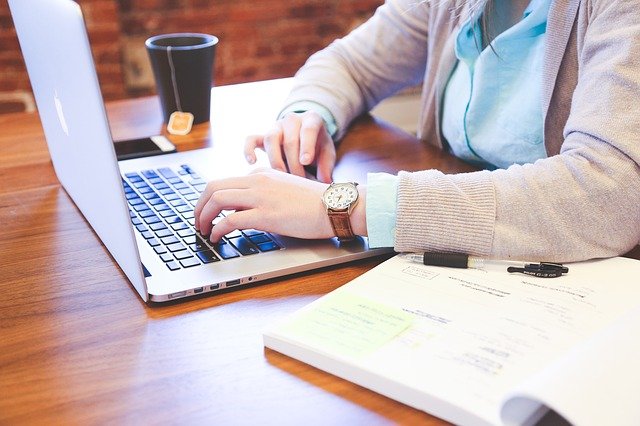 zbliżenie na ręce kobiety pracującej przy laptopie, obok otwarty notes i kubek z herbatą