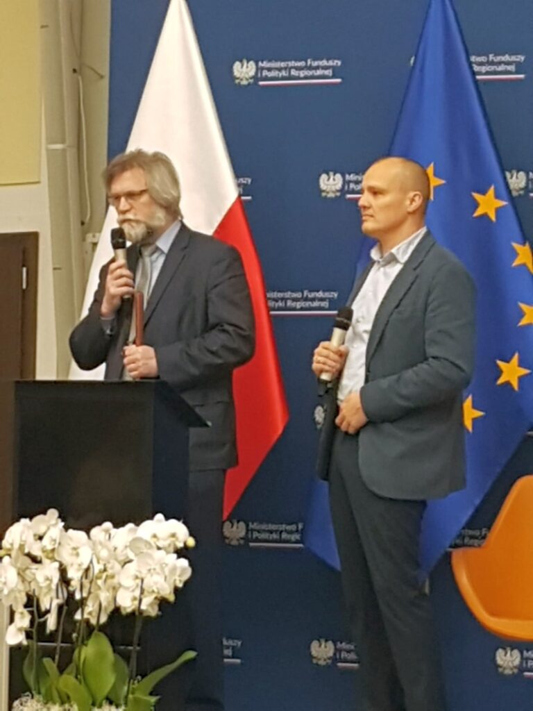 Dwóch mężczyzn stojących z mikrofonami przy mównicy, w tle flaga Polski i Unii Europejskiej