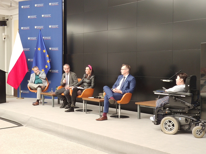 Pięć osób siedzących, w tym jedna na wózku inwalidzkim podczas rozmowy, w tle flaga Polski i Unii Europejskiej