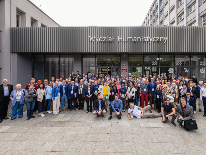 Grupa osób pozuje do zdjęcia. W tle budynek z napisem Wydział Humanistyczny