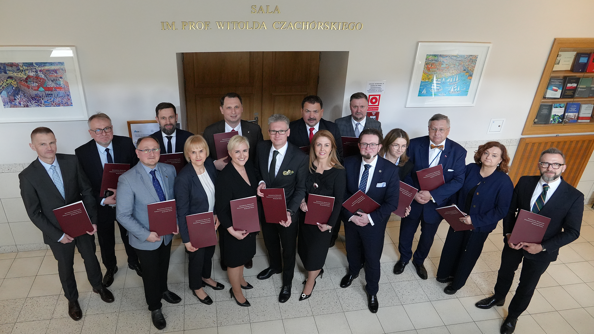 Fotografia przedstawia 16 Dziekanów Wydziałów Prawa, którzy wspólnie pozują do zdjęcia. Każde z Państwa ma w ręku teczkę z napisem Konferencja Dziekanów Publicznych Uniwersyteckich Wydziałów Prawa