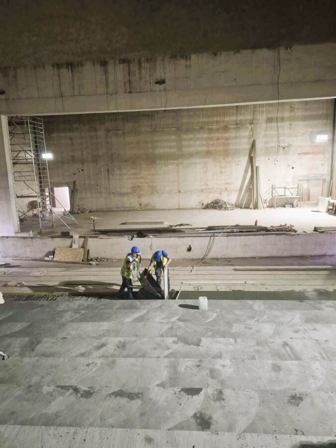 Rewitalizowana aula od strony widowni. Pracownicy wylewają beton pod nowe stopnie widowni. W głębi widoczne są scena i rusztowanie.