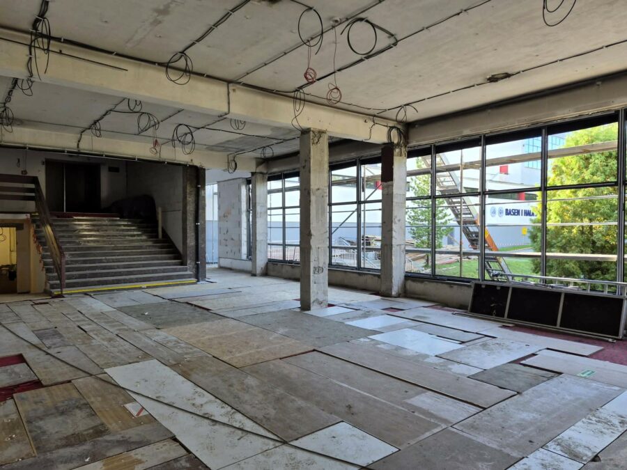 Wnętrze przyszłego foyer w stanie surowym. Widoczne są betonowe kolumny, duże okna, klatka schodowa oraz prowadzone instalacje elektryczne.