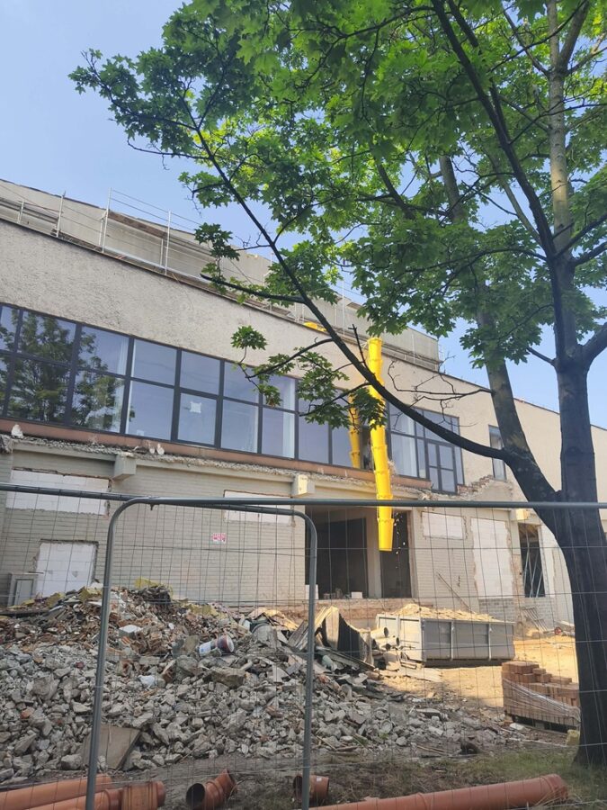 Fot. 1 Za ogrodzeniem trwa remont budynku, wokół którego ustawiono rusztowania i składowane są materiały budowlane. Na pierwszym planie znajduje się tablica informacyjna o dofinansowaniu inwestycji z Funduszy Europejskich dla projektu Uniwersytetu Śląskiego. Nad placem budowy unosi się pył.