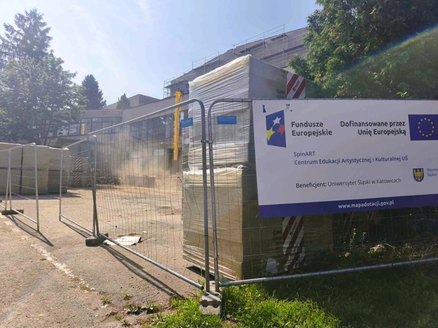 Fot.3 Budynek w trakcie przebudowy, otoczony tymczasowym ogrodzeniem placu budowy. Przed budynkiem znajdują się hałdy gruzu, kontener na odpady i materiały budowlane, a nad wejściem zamontowano rękaw do zrzutu gruzu. Górna część budynku jest osłonięta rusztowaniami, a elewacja pozostaje w trakcie prac.
