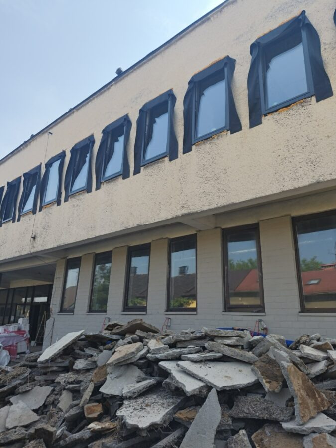 Fot.4 Na zdjęciu widać elewację budynku w trakcie przebudowy. Na pierwszym planie znajduje się sterta gruzu z fragmentów betonowych płyt i innych materiałów rozbiórkowych. Nowe okna na górnej kondygnacji są zabezpieczone czarną folią ochronną, a przy wejściu widoczne są materiały budowlane przygotowane do dalszych prac.