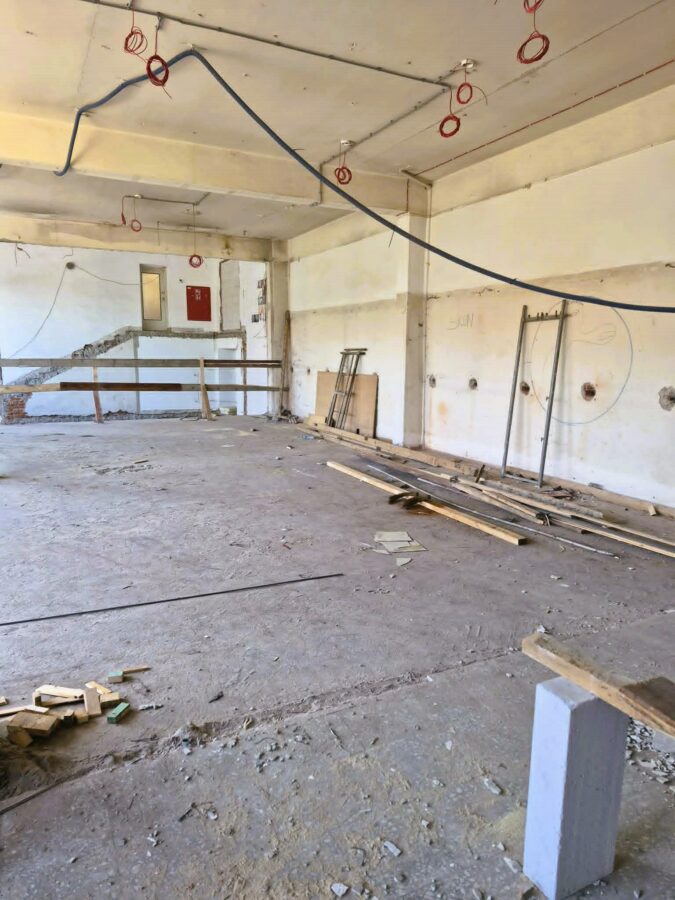 8. Zdjęcie przedstawia wnętrze dawnej sali baletowej w trakcie remontu. Widoczne są jasne ściany i słupy konstrukcyjne ze śladami prowadzonych prac. Pod sufitem ciągną się odsłonięte, nowe przewody instalacji elektrycznej. Na podłodze znajdują się elementy konstrukcyjne, metalowe rury, gruz i drewniane deski. Po prawej stronie o ścianę oparto kilka drabin, a w pobliżu przeszklonych drzwi leży kolejna. W tle widoczne są przejścia do sąsiednich pomieszczeń oraz puste miejsce po zdemontowanych schodach.