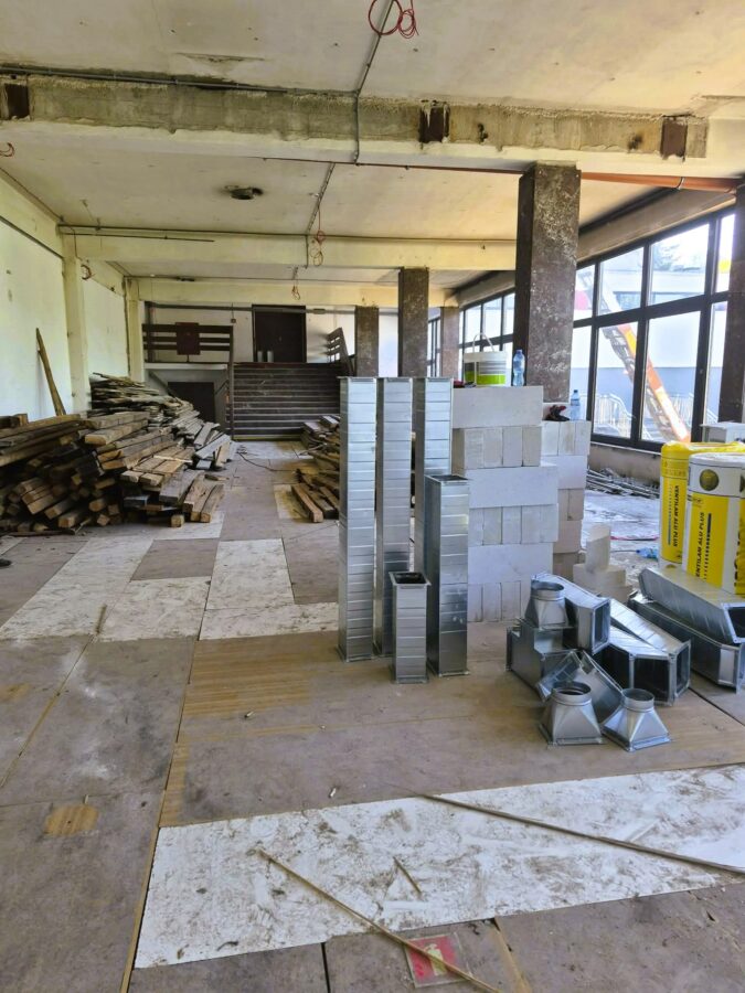 9. Zdjęcie przedstawia foyer w trakcie remontu. We wnętrzu znajdują się stosy drewnianych belek, bloczków oraz metalowych elementów instalacji wentylacyjnej. Posadzka jest zabezpieczona drewnianymi płytami, na których widoczne są gruz i pozostałości po pracach. Po prawej stronie znajdują się duże okna, a w tle schody i drzwi prowadzące do dalszej części budynku.